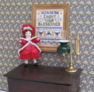 A tiny doll's house doll's doll, complete with her pleated bonnet, standing next to the 'Count Your Blessings' sampler