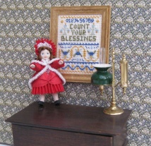 A tiny doll's house doll's doll, complete with her pleated bonnet, standing next to the 'Count Your Blessings' sampler